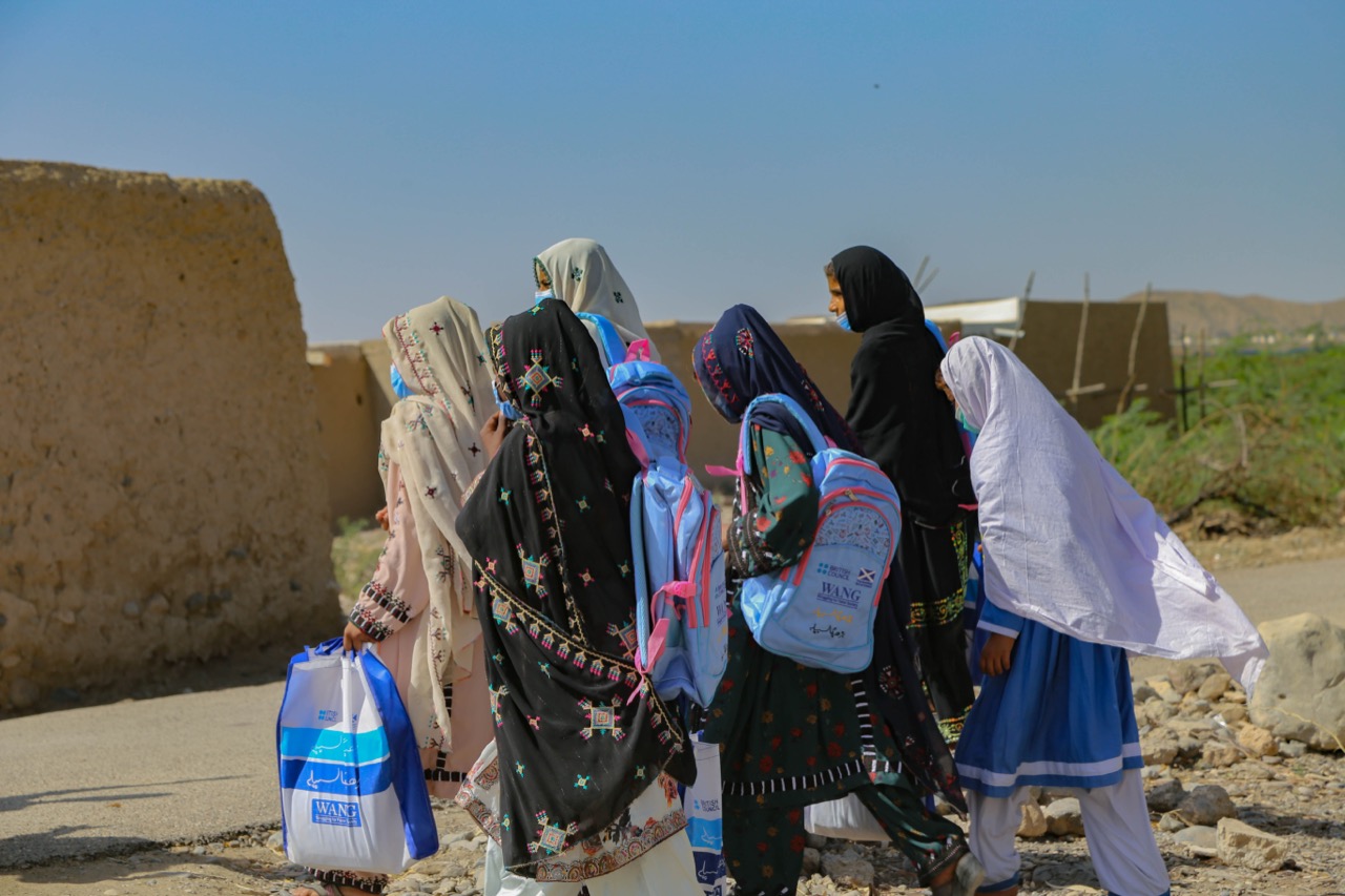Girls with school bags walking on a dusty rural road in Balochistan with mountains behind